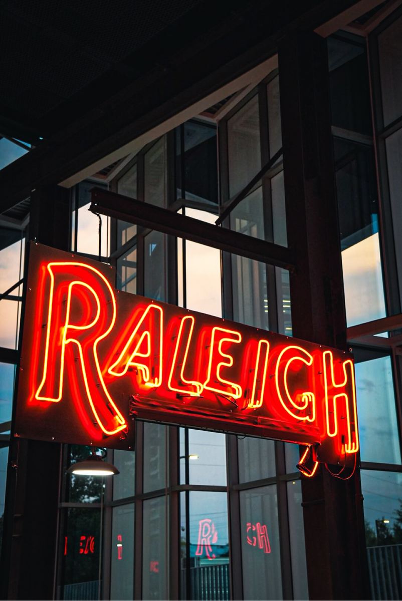 Red neon 'Raleigh' sign glowing in a downtown storefront window at dusk