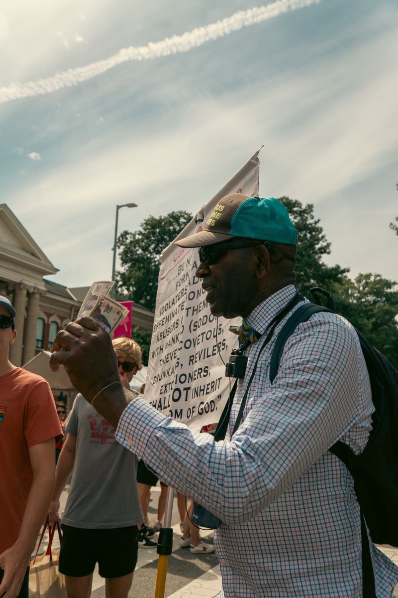 Man in sunglasses and cap holding a sign at a daytime street festival in Raleigh